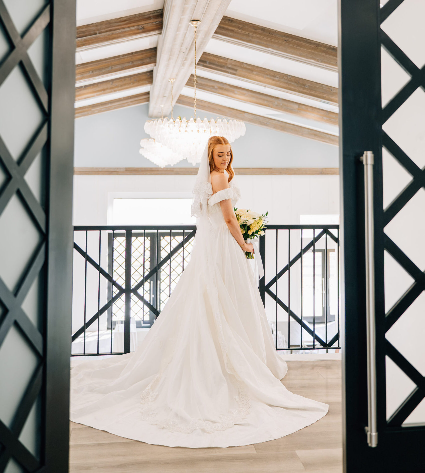 A bride in a white wedding dress stands on the dark wood and wrought-iron balcony at Riverbridge Event Center, looking out over a grand ballroom with high ceilings, exposed wood beams, and large sparkling chandeliers in Spanish Fork, Utah.