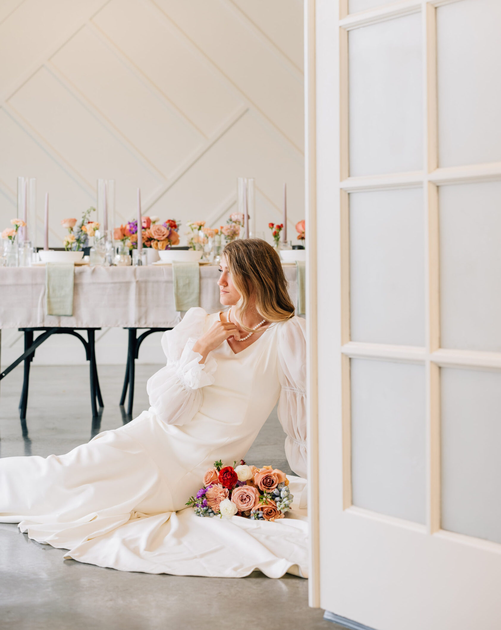A bride in a sleek modern wedding dress sits at a sweetheart table decorated with geometric gold centerpieces and greenery inside the large ballroom with high ceilings at The Azalea wedding venue in South Jordan, Utah.